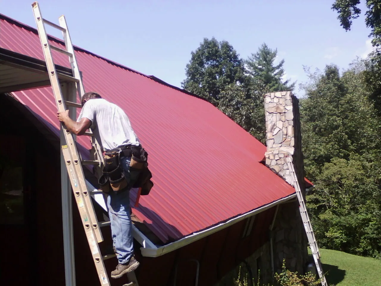 Roofer on a ladder beside a red metal roof and stone chimney — Soffit & Fascia Repair by Roofing Brothers in Oak Park